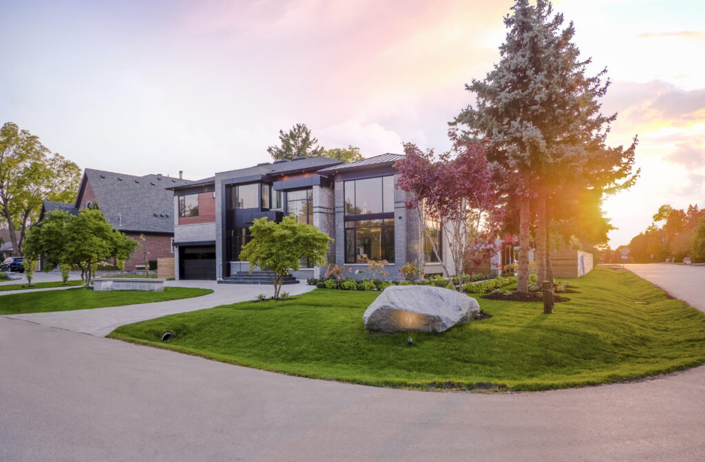 Modern two-story house with large windows, a landscaped front yard, trees, and a boulder, set on a corner lot with a driveway, at sunset with a warm glow in the sky.