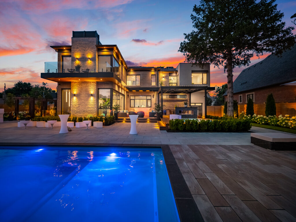 Modern two-story house with large windows and outdoor lighting at sunset, featuring a landscaped yard, illuminated swimming pool, and patio with contemporary furniture.