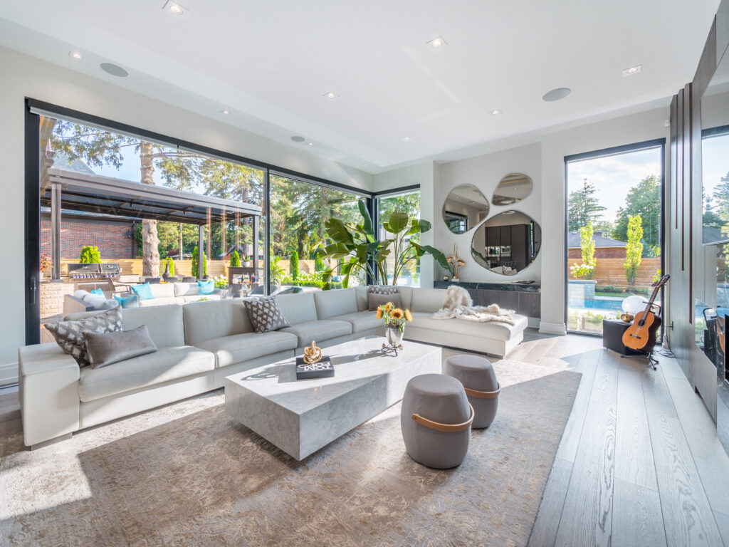 A modern, bright living room with large windows, a white sectional sofa, a marble coffee table, decorative mirrors, indoor plants, and a view of a patio and pool outside. A guitar sits near the TV on the right.