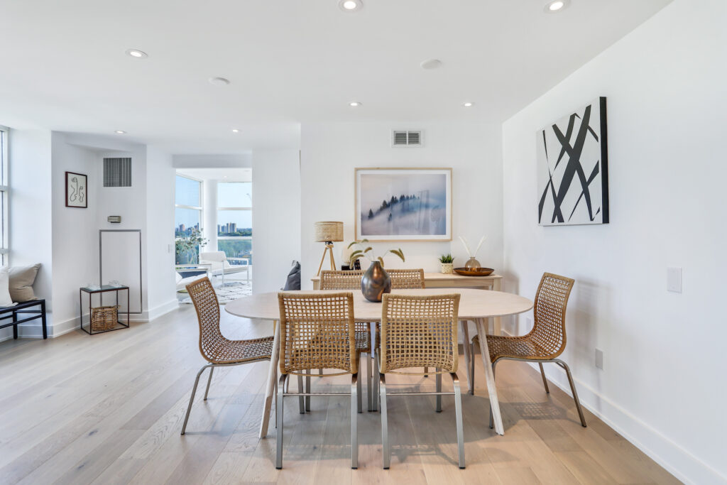 A modern dining room with a light wood table, six woven chairs, minimalist decor, light wood flooring, and white walls. Large windows and framed artwork are visible, creating a bright and airy atmosphere.