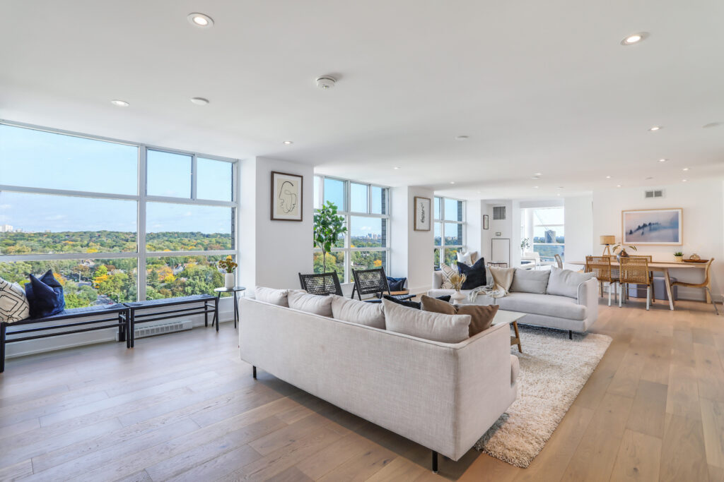 Bright, modern living and dining area in a downtown condo renovation with open-concept layout. Large windows, light hardwood floors, neutral sofas, and scenic treetop views create a cozy yet elegant feel accented by minimalist decor and lush plants.