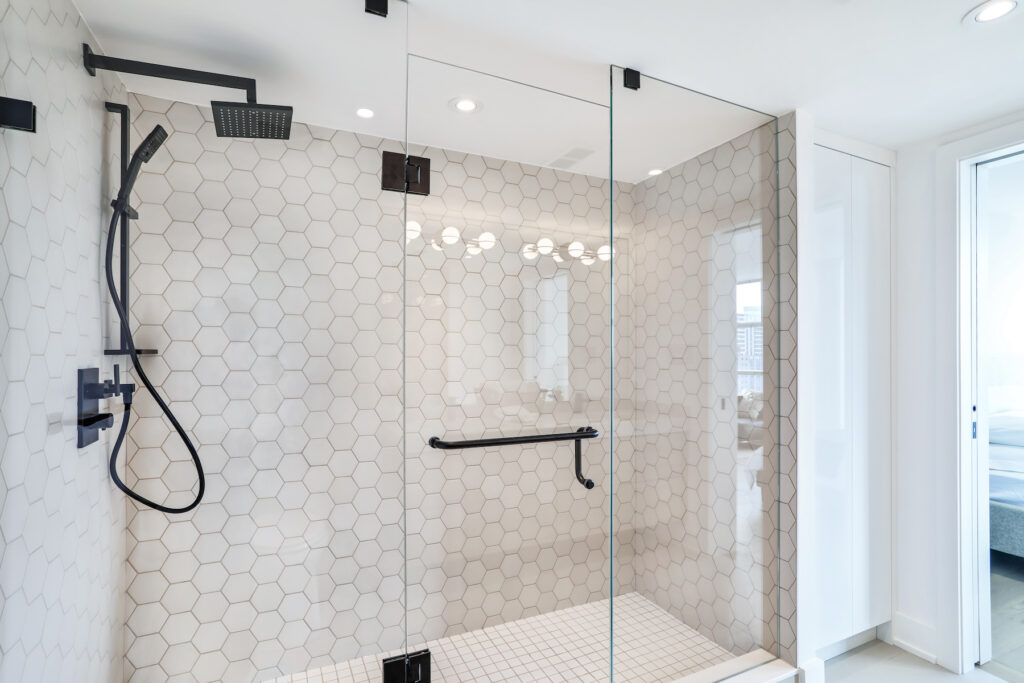 Modern bathroom shower with clear glass doors, black fixtures, a rain showerhead, handheld shower, and white hexagonal tile walls. A grab bar is mounted on the glass, and natural light fills the space.