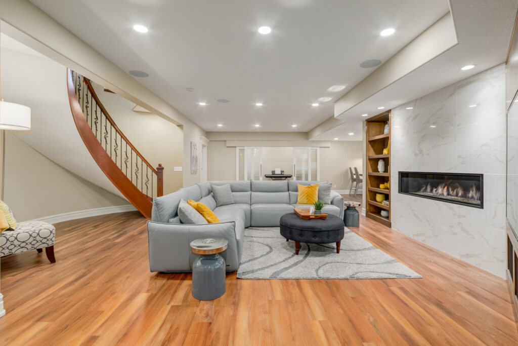 Modern living room with wood floors, a light-gray sectional sofa, round ottoman, patterned armchair, built-in shelves, and a sleek fireplace set in a white marble wall. Curved staircase visible in the background.