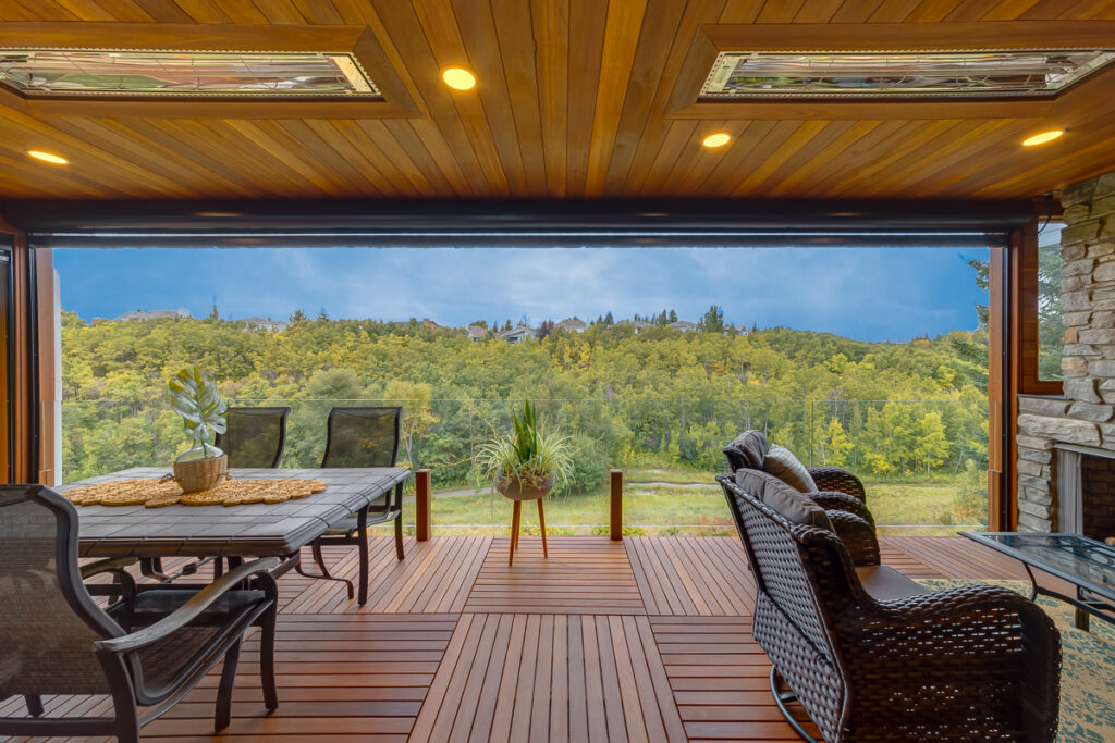 A covered patio with wooden flooring, outdoor dining and lounge furniture, overlooks a lush green forest and distant hills under a cloudy sky.