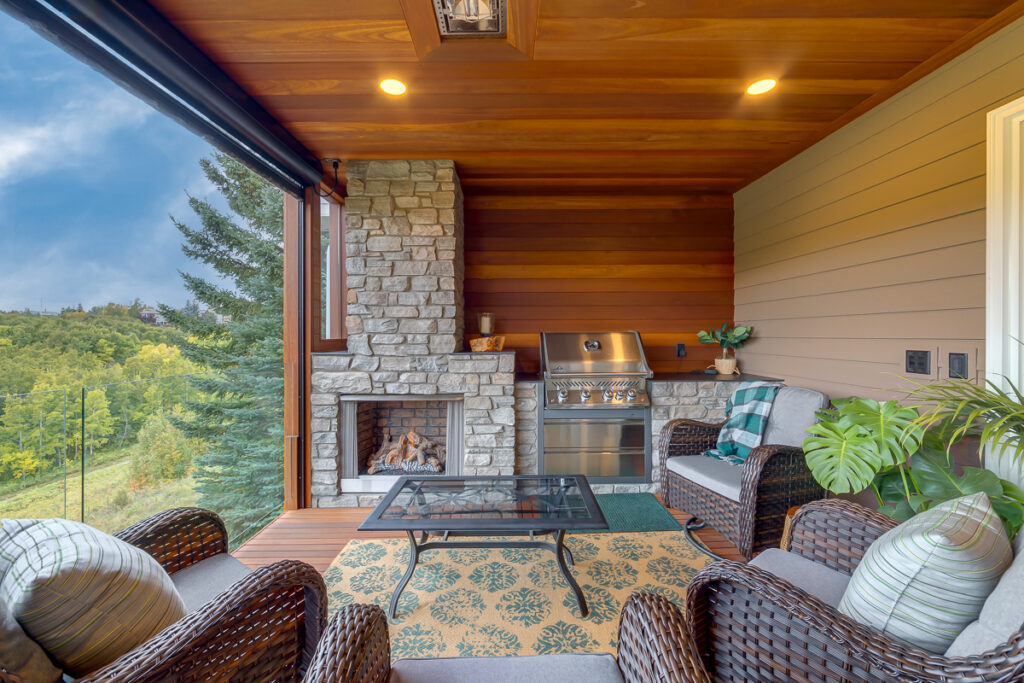 A cozy covered patio with wicker chairs, cushions, a rug, a stone fireplace, and a stainless steel grill. Large open windows reveal a scenic, green outdoor view. Warm wood and stone accents create an inviting atmosphere.