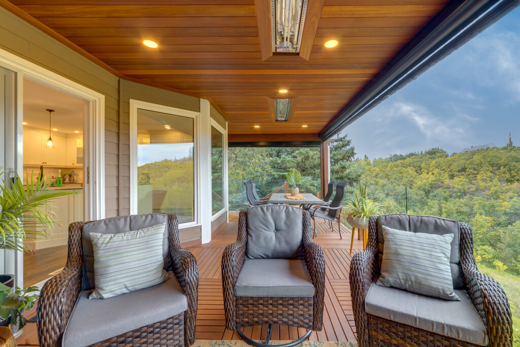 A covered deck with wicker chairs, gray cushions, and pillows facing a dining table. The deck overlooks lush green trees and has large glass doors connecting to a modern kitchen indoors.