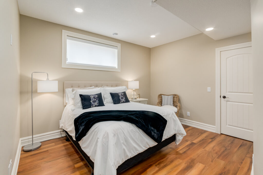 A cozy bedroom with a double bed, white bedding, navy throw pillows, and a dark blue blanket. There are two bedside lamps, a small side table, a striped armchair, and wood flooring. A window is above the bed.