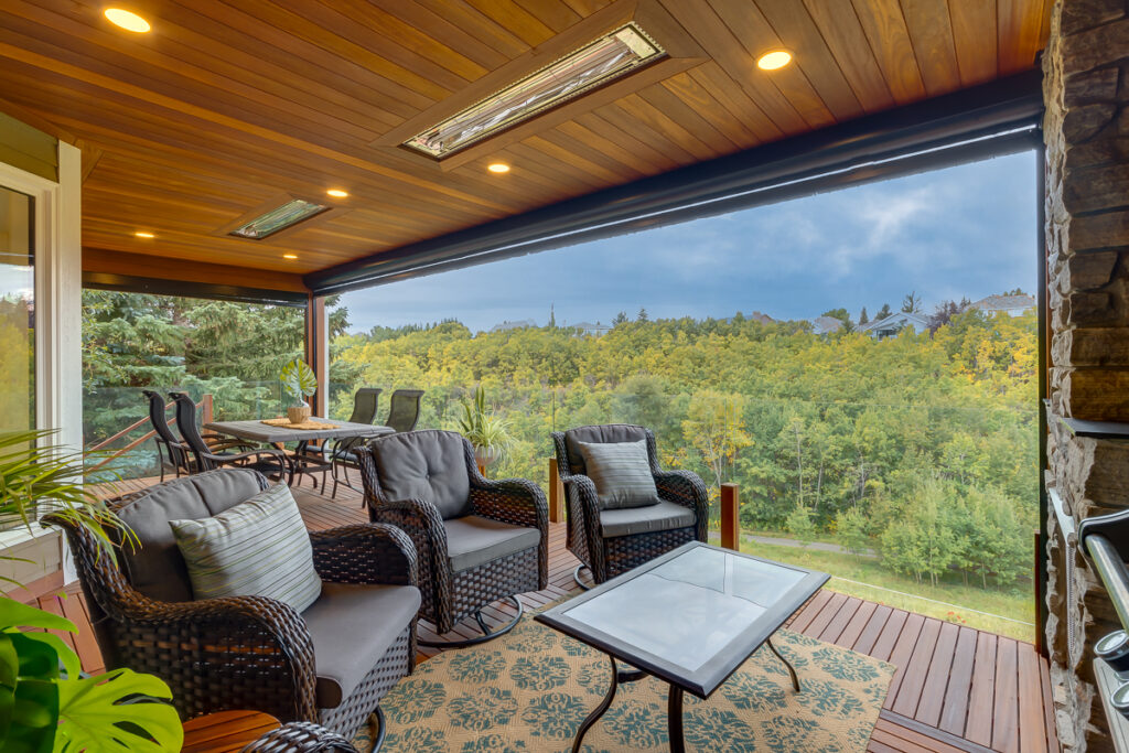 A covered wooden deck overlooks a lush green forest. The deck is furnished with cushioned wicker chairs, a table, a dining set, and features recessed lighting in the wooden ceiling and a decorative rug under the seating area.
