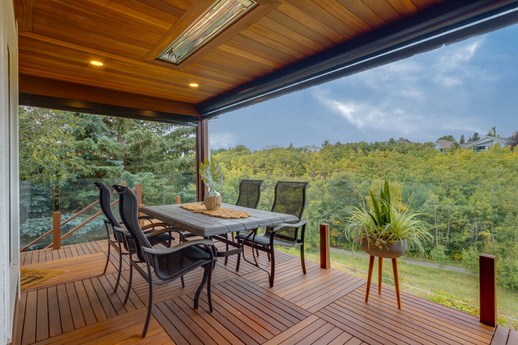A covered wooden patio with a dining table and six chairs overlooks a lush, green forest. Potted plants decorate the space, and recessed ceiling lights brighten the area. The patio has glass railings and a scenic view.