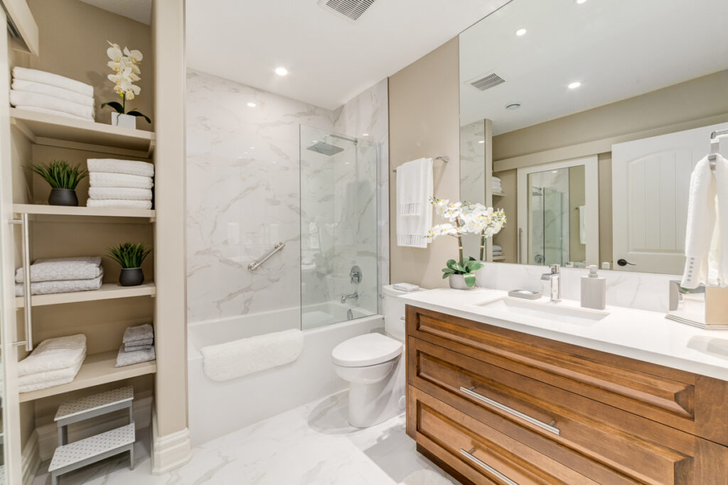 Modern bathroom with a glass-enclosed bathtub, marble tiles, wooden vanity, large mirror, stacked towels on shelves, and white orchids on the counter, creating a clean and elegant look.