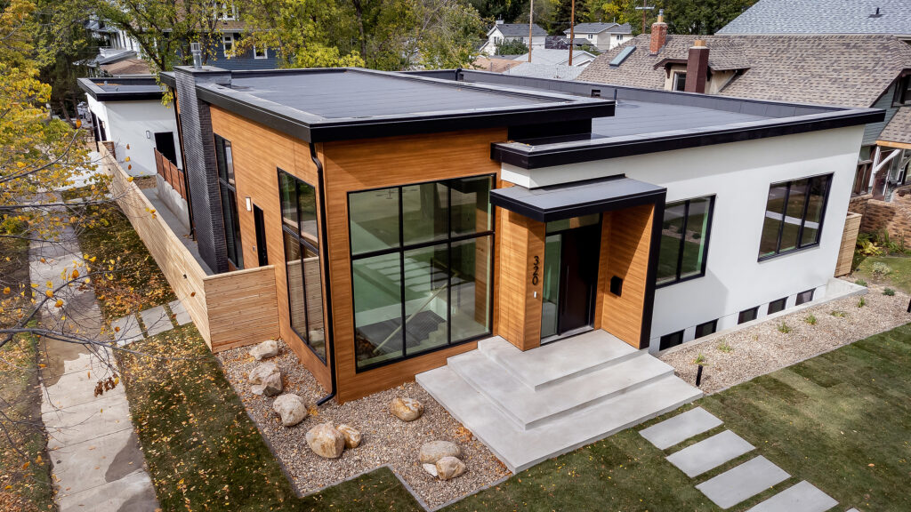 A modern house with flat roofs, large floor-to-ceiling windows, wood and white exterior, concrete steps, and minimalist landscaping with rocks and grass, seen from an elevated angle.