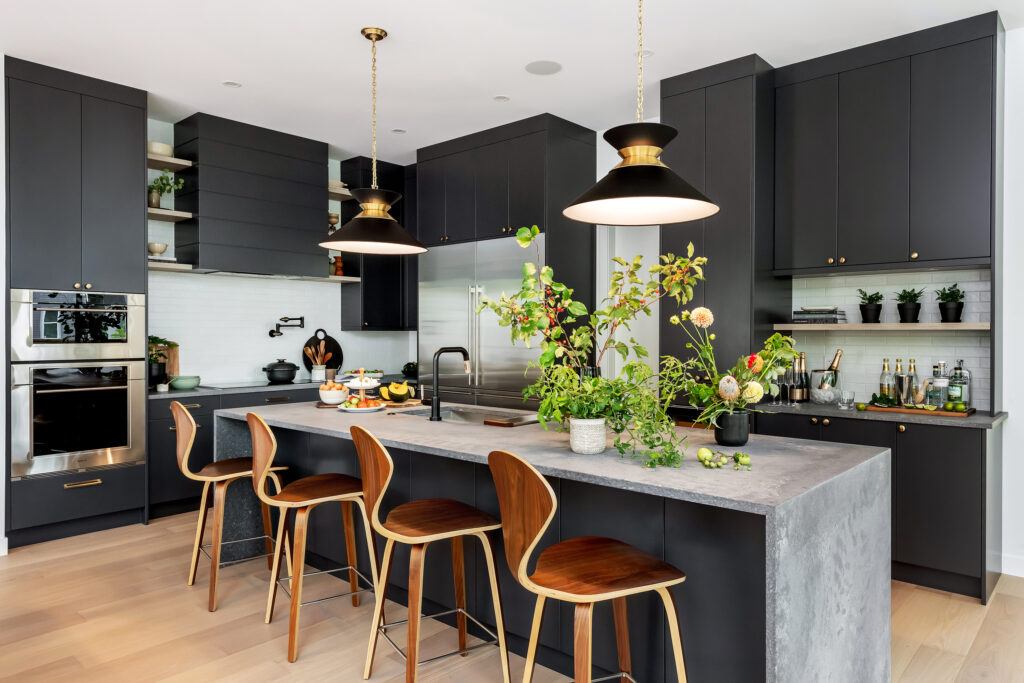 Modern kitchen with matte black cabinets, a concrete island with wooden stools, brass pendant lights, and greenery on the countertop. Open shelves display dishes and drinks, and stainless steel appliances are integrated.