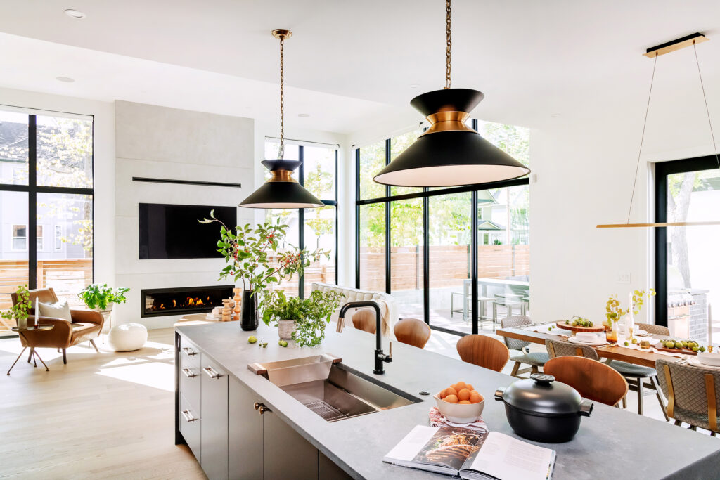 Modern, bright kitchen with large windows, island with sink, black pendant lights, and an open cookbook. Dining table is set, and a living area with chairs and a fireplace is visible in the background.