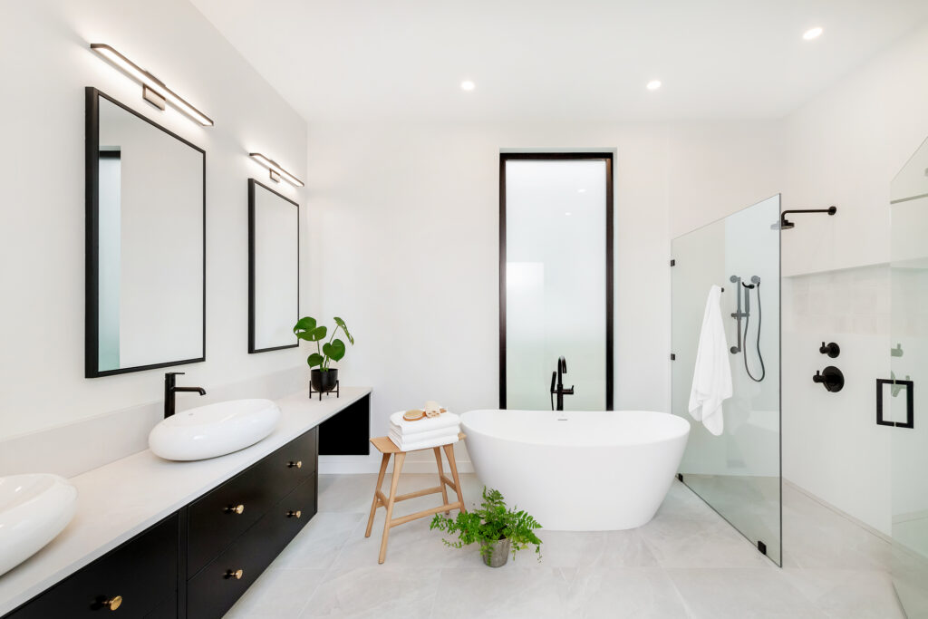 Modern bathroom with double sinks, black fixtures, a freestanding white bathtub, glass shower, two large mirrors, a wooden stool with towels, a potted plant on the floor, and bright white walls.
