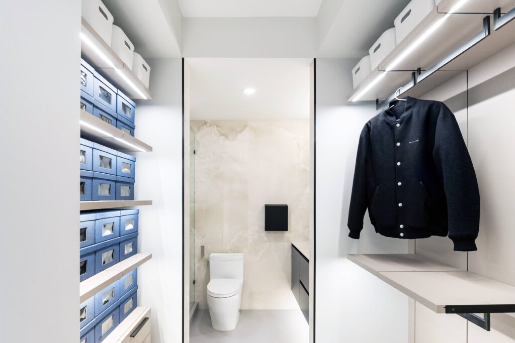 A modern walk-in closet with shelves of blue and white storage boxes leads to a small bathroom with a toilet and minimalist decor; a black jacket hangs on the right.
