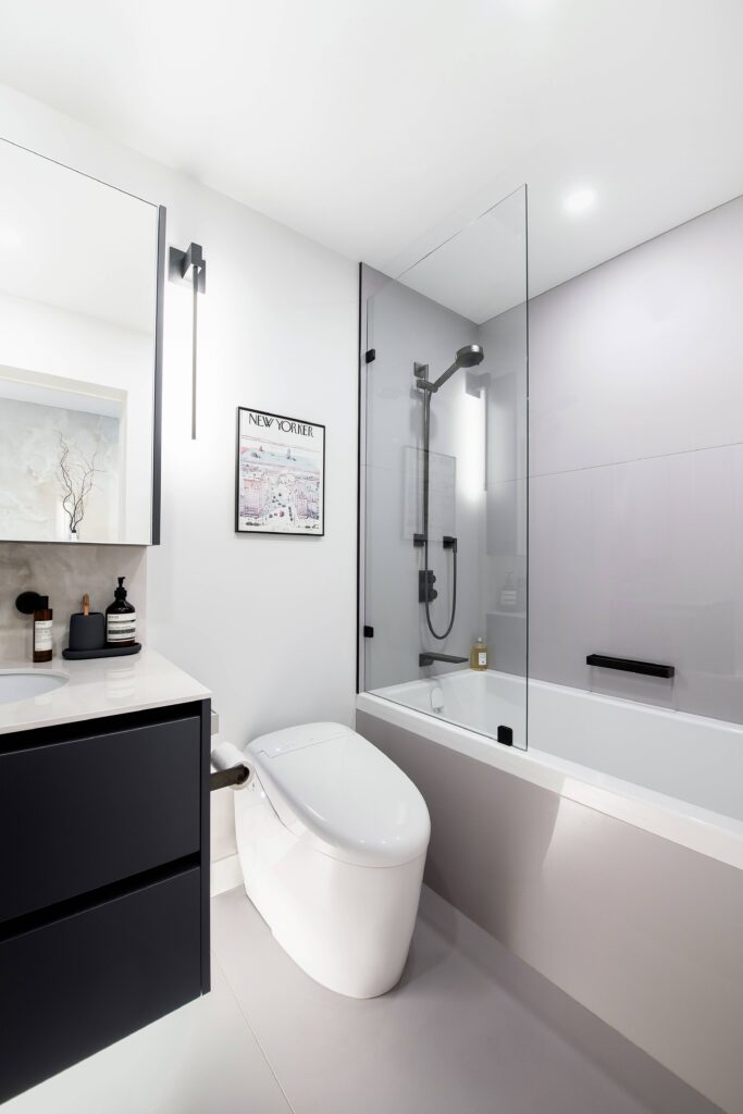 Modern bathroom with a sleek white toilet, black vanity, glass-enclosed bathtub with shower, wall-mounted lighting, and neutral decor. Minimalist design with clean lines and light-colored walls.
