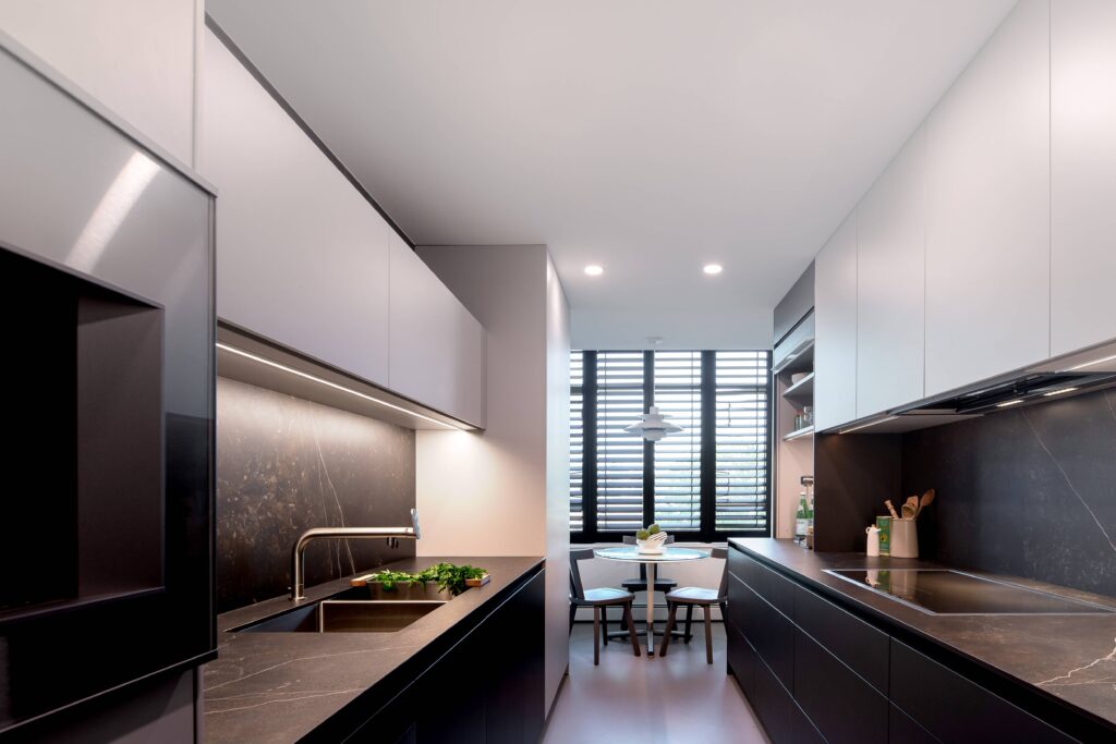 Modern kitchen with sleek black countertops and cabinets, built-in appliances, and under-cabinet lighting. A small dining area with a round table and two chairs is by large windows with horizontal blinds.
