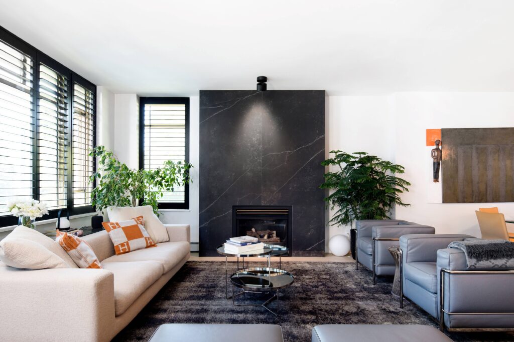 Modern living room with a beige sofa, gray armchairs, glass coffee tables, a large black stone fireplace, indoor plants, and contemporary art on the wall. Large windows with shutters let in natural light.