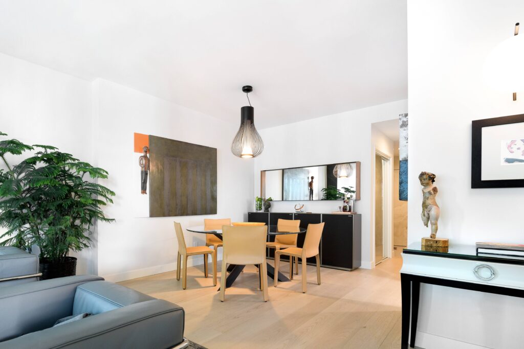 Modern dining area with a round wooden table, four light tan chairs, a dark sideboard, pendant light, indoor plant, wall art, and decorative sculptures in a bright room with light wood floors.