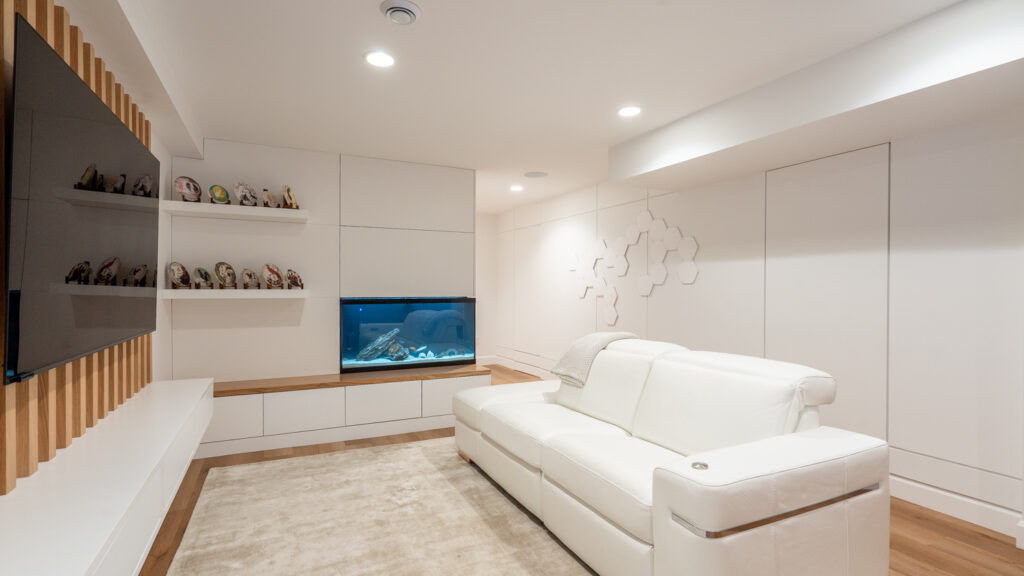 A modern living room with white walls and furniture, a large flat-screen TV, wall shelves with small figurines, an aquarium built into the wall, and soft recessed lighting.