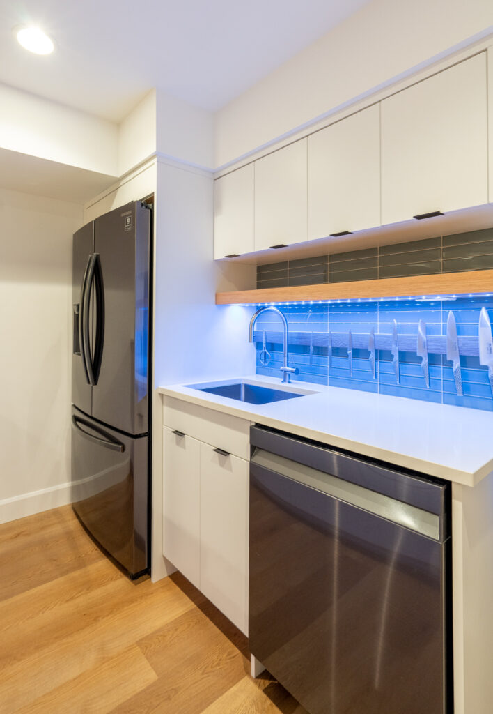 Modern kitchen with white cabinets, stainless steel refrigerator and dishwasher, wood flooring, and a blue LED light strip under the upper cabinets above the sink.