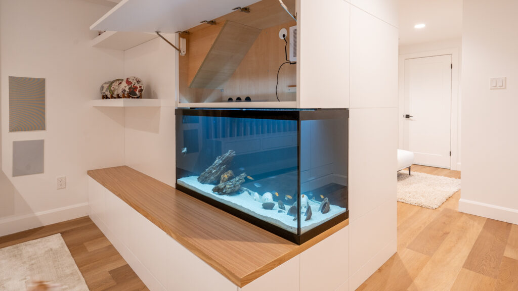 A modern room features a built-in fish tank with clear water, rocks, and fish, set in a white cabinet with wood accents, adjacent to a hallway with light wood floors and white walls.
