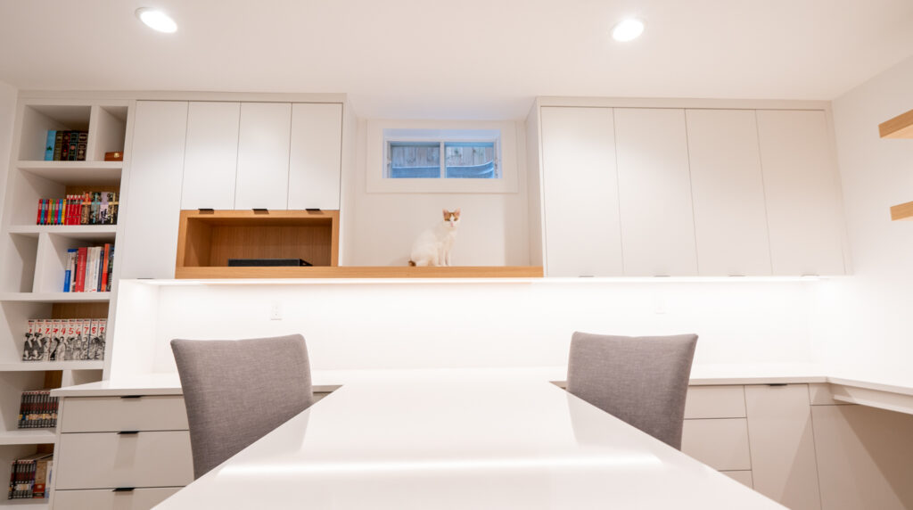 A white and orange cat sits on a wooden shelf in a modern, bright home office with white cabinets, bookshelves with books and figurines, and two gray chairs at a white desk.