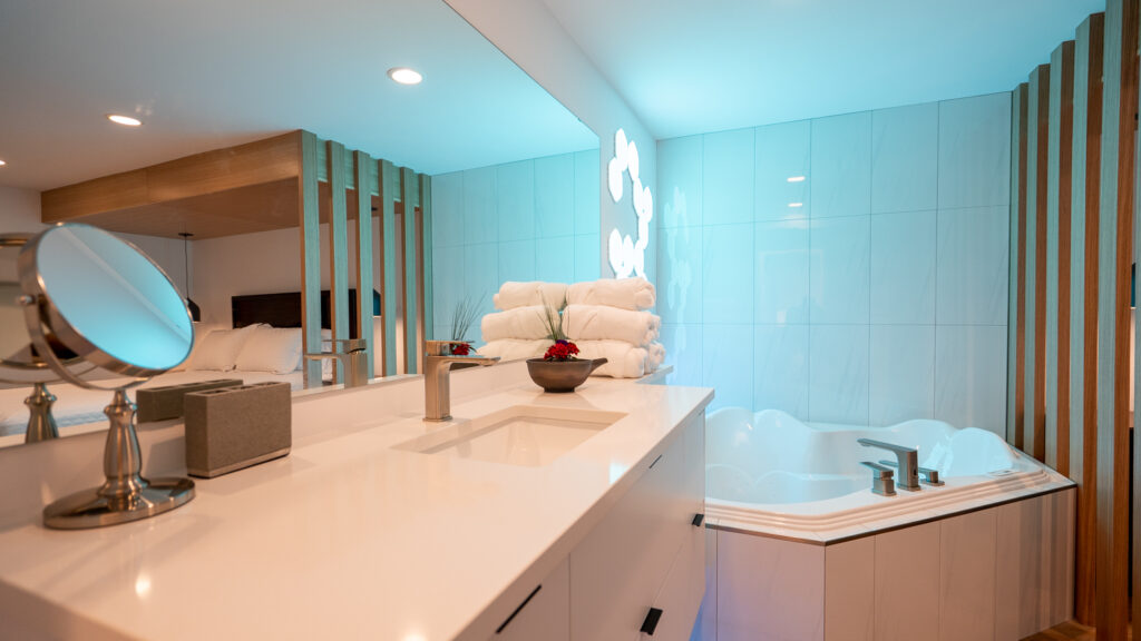 Modern bathroom with a white countertop, mounted faucet, mirror, stacked towels, decorative bowl, and a built-in corner bathtub with blue accent lighting and wooden slats as partial dividers.