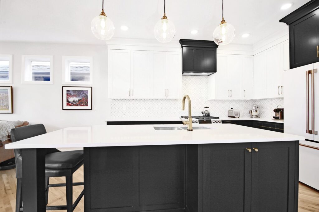 Modern kitchen with white cabinets, black island, gold faucet, three pendant lights, stove, fridge, and barstool. Art hangs on the wall and natural light enters through small windows.