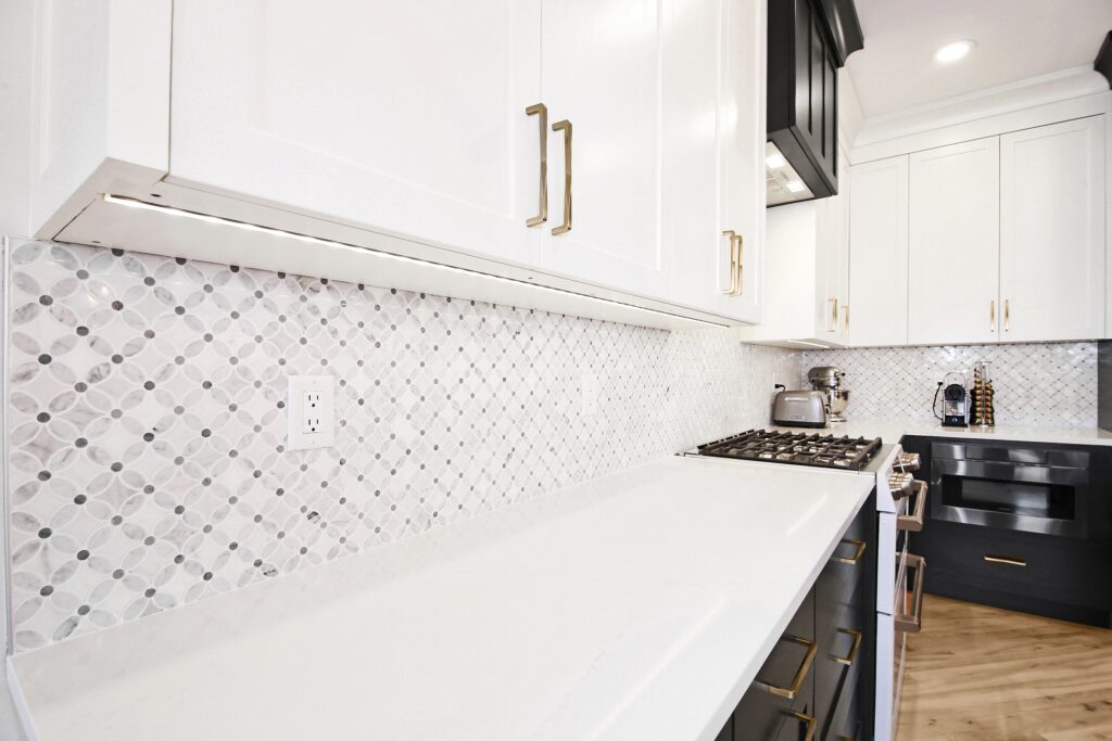 Modern kitchen with white upper cabinets, gold handles, a geometric patterned backsplash, white countertop, a gas stove, toaster, coffee maker, and wooden floor. The space has a clean and bright appearance.