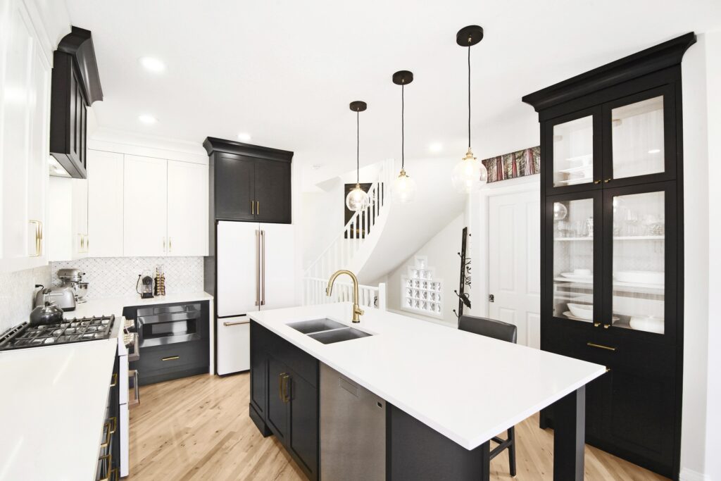 Modern kitchen with black and white cabinets, a white island with a gold faucet, pendant lights, wooden floor, and white tile backsplash. Stairs and a white display shelf are visible in the background.