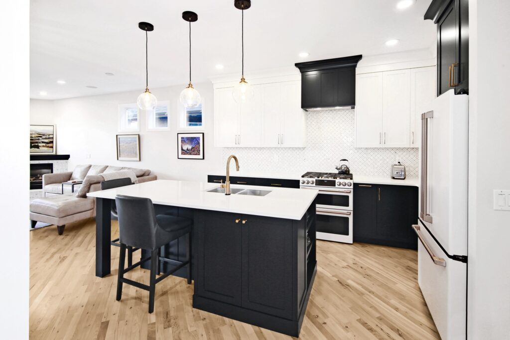 Modern kitchen with white cabinets, black island with sink, gold faucet, pendant lights, light wood floors, and a nearby living area with a beige sectional sofa and wall art.
