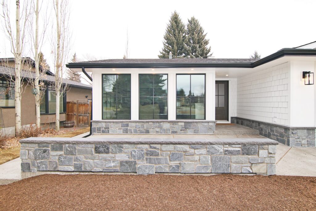 Modern house exterior with large windows, stone accents on the lower wall, light siding, and a covered front porch. There are trees and neighboring homes visible in the background.