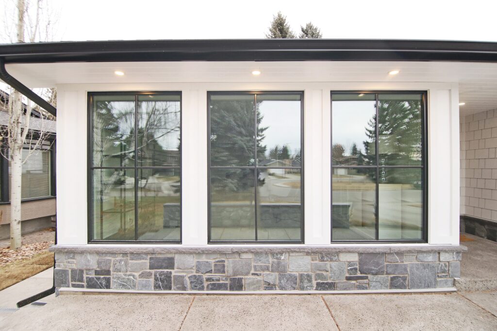 A modern house exterior featuring three large grid-style windows framed in white with a stone base, set under a white overhang with recessed lighting. Trees and a street are visible through the glass.
