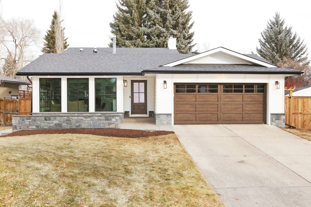 A modern single-story house with large front windows, stone accents, and a two-car garage with wooden doors. The driveway and lawn are in front, with trees and a wooden fence in the background.