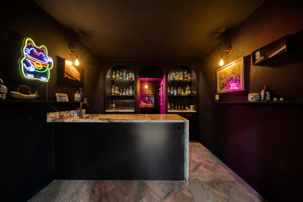 A small, modern bar with dark walls, neon lighting, and a marble counter. Shelves behind the bar display various bottles. Neon cat sign and colorful framed art decorate the walls, creating a cozy, vibrant atmosphere.