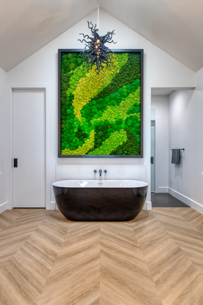 Modern bathroom with a black-and-white freestanding tub, wood-patterned chevron flooring, large green moss wall art above the tub, and a unique ceiling light fixture resembling twisting branches.