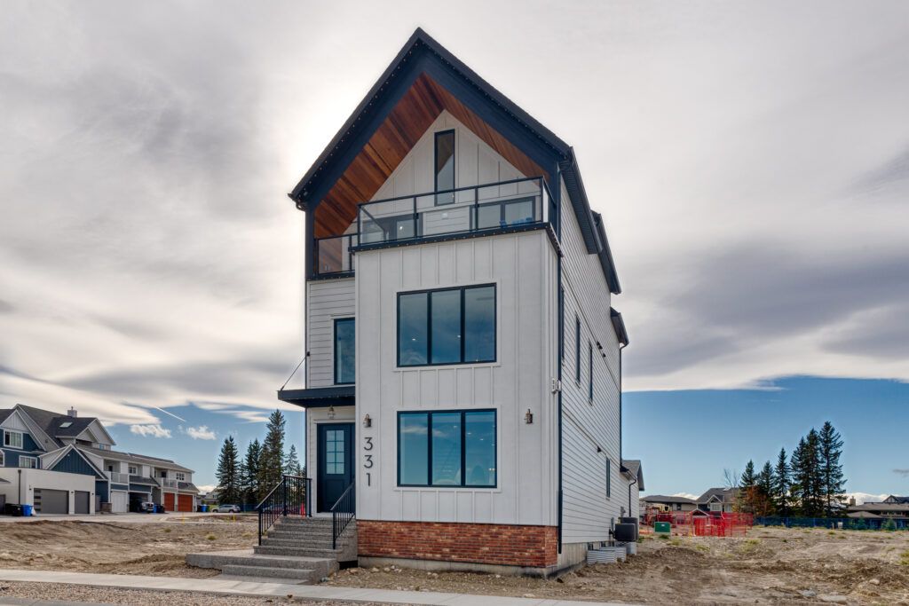 A modern three-story house with white siding, large windows, and a front balcony, located on a dirt lot in a suburban neighborhood under a cloudy sky. The house number 331 is visible.