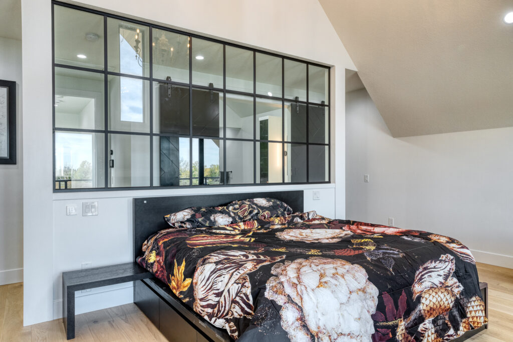 A modern bedroom with a bed featuring a bold, floral-patterned comforter. Behind the bed is a large window with black grid frames, showcasing another room and natural light. The walls are white with light wood flooring.