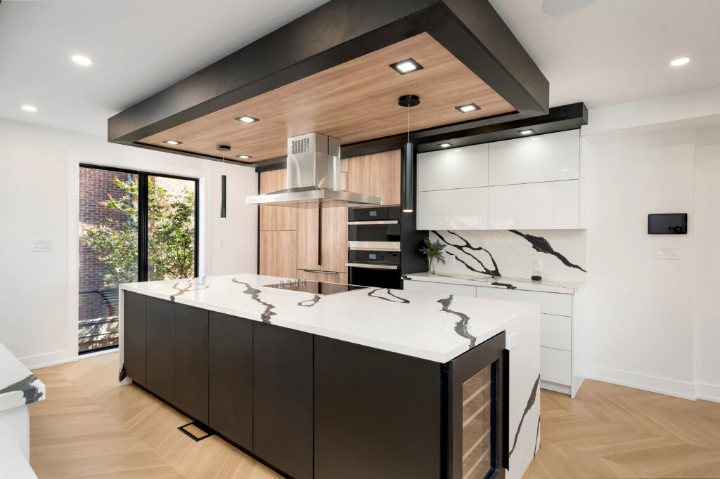 Modern kitchen with a large black and white island, marble countertops, built-in wine fridge, wood flooring, sleek cabinetry, a stainless steel range hood, and large window letting in natural light.