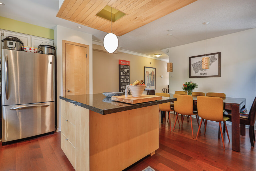 kitchen with a wooden island, black countertop, stainless steel fridge, and a dining area with six orange chairs and a wooden table. Pendant lights hang above both the island and dining table.