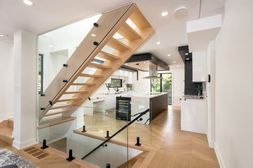 Modern, open-concept kitchen with wood and glass staircase, white cabinetry, large windows, light wood floors, and sleek black and stainless steel accents. Natural light fills the space, creating a bright, airy atmosphere.