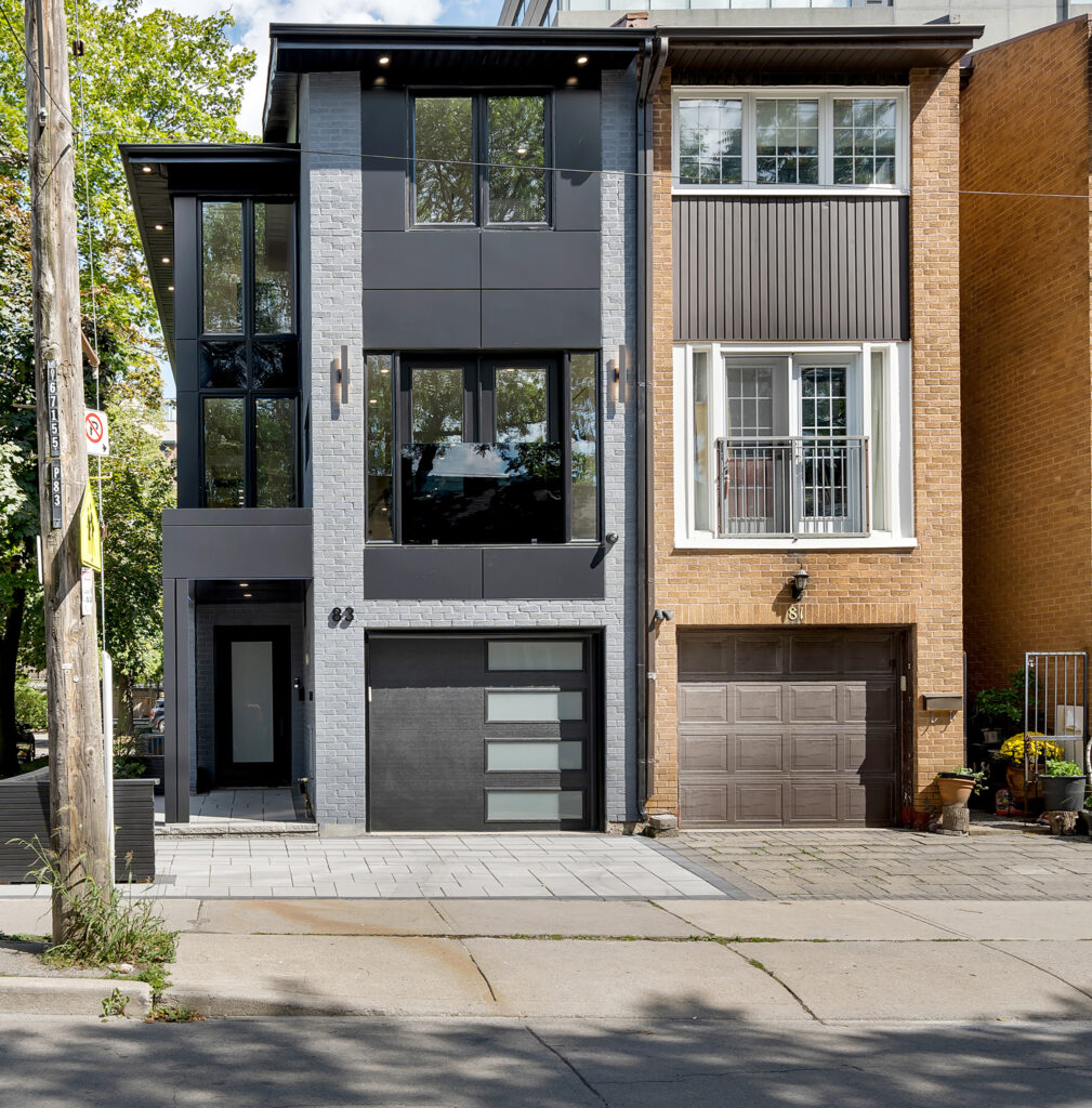 Modern black and gray townhouse with large windows and glass garage door, next to a traditional brown brick house with a metal balcony and brown garage door. Trees and sidewalk are visible in front.
