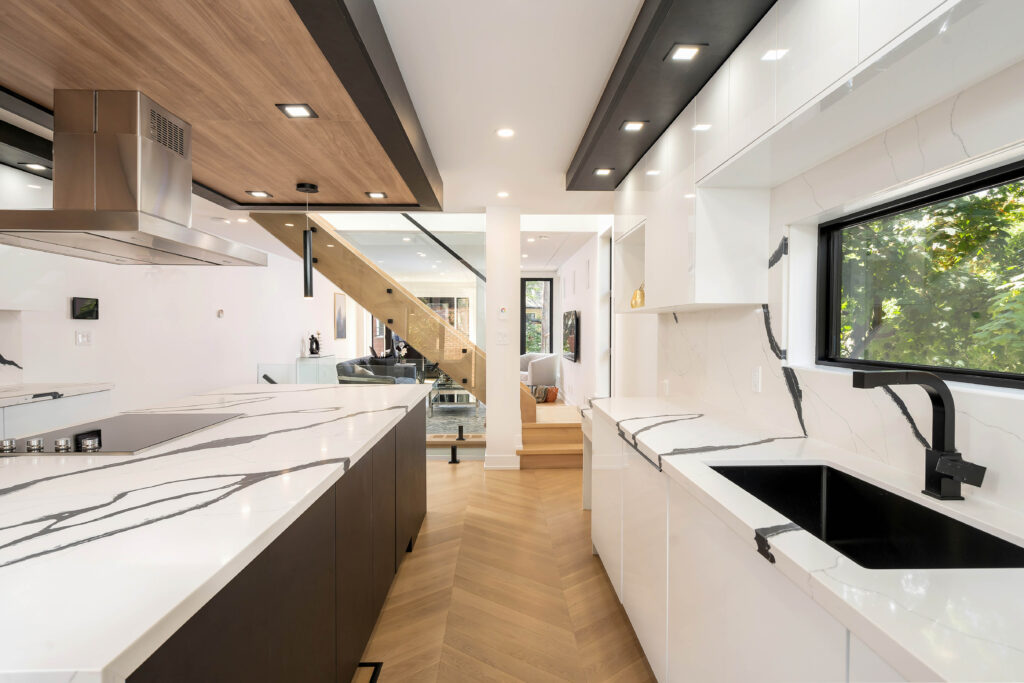 Modern kitchen with white marble countertops, black fixtures, light wood floors, and a wood-accented ceiling. Large window above the sink lets in natural light. Open stairs and living area are visible beyond the kitchen.
