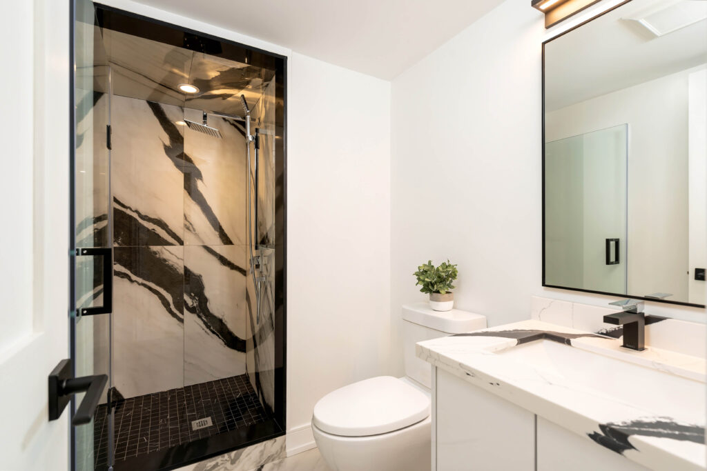 Modern bathroom with white walls, a black and white marble shower, a white toilet, a vanity with a marble countertop, a rectangular mirror, and a small green plant on the counter.