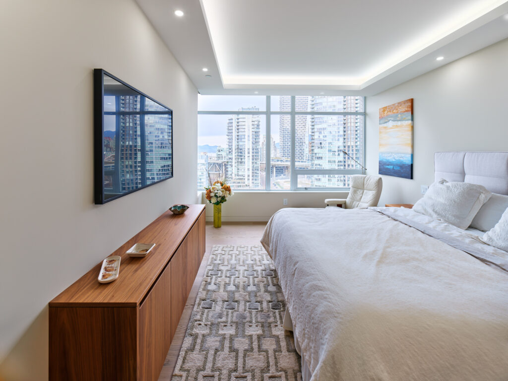 A modern bedroom with a large bed, white bedding, a wooden sideboard, wall-mounted TV, armchair, and large windows offering a city skyline view. A vase with flowers and a framed painting are also visible.