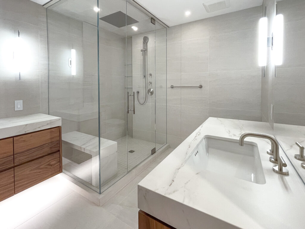 Modern bathroom with a glass-enclosed shower, bench seating, rainfall showerhead, wood cabinets, and a white marble countertop with a built-in sink and brushed metal fixtures. Bright lighting and neutral tile throughout.