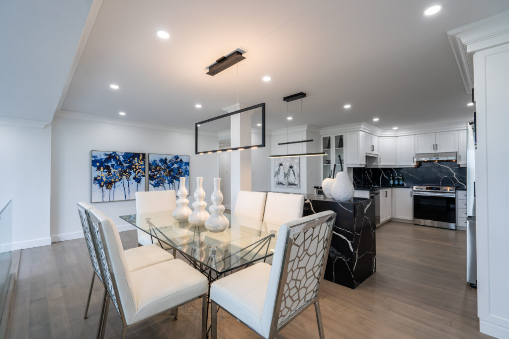 Modern dining area with glass table, white upholstered chairs, and decorative vases. Open kitchen with white cabinets, black marble island, and stainless steel appliances. Abstract artwork on the wall and recessed lighting throughout.