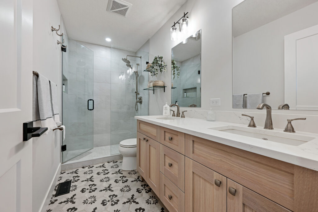 Modern bathroom with light wood double vanity, white countertop, large mirror, floral-patterned tile floor, glass-enclosed shower, wall-mounted lights, silver fixtures, and neatly hung towels.