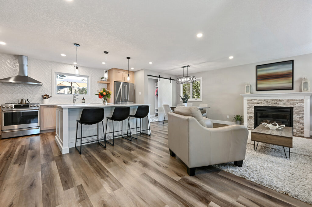 Modern open-concept kitchen and living room with light wood floors, white cabinets, a breakfast bar with three black stools, a cozy seating area, and a fireplace with a large framed art piece above it.